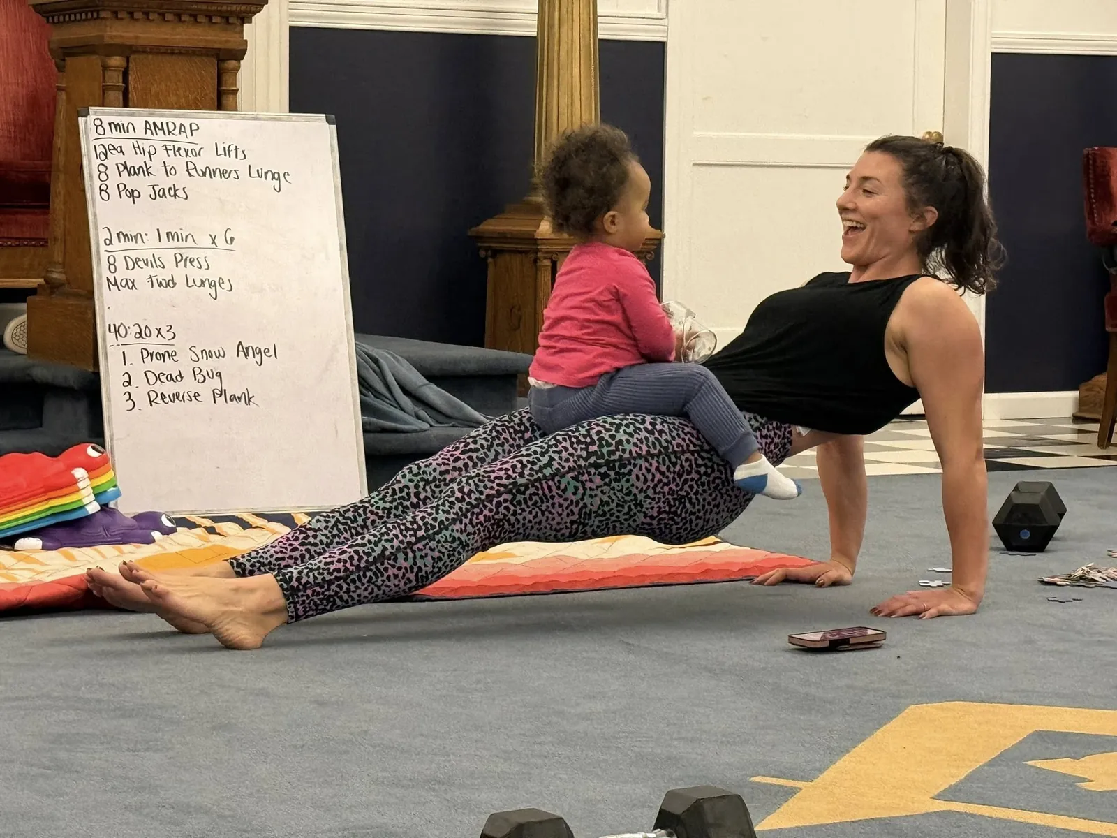 Member holding a plank with her kid climbing on her back during class