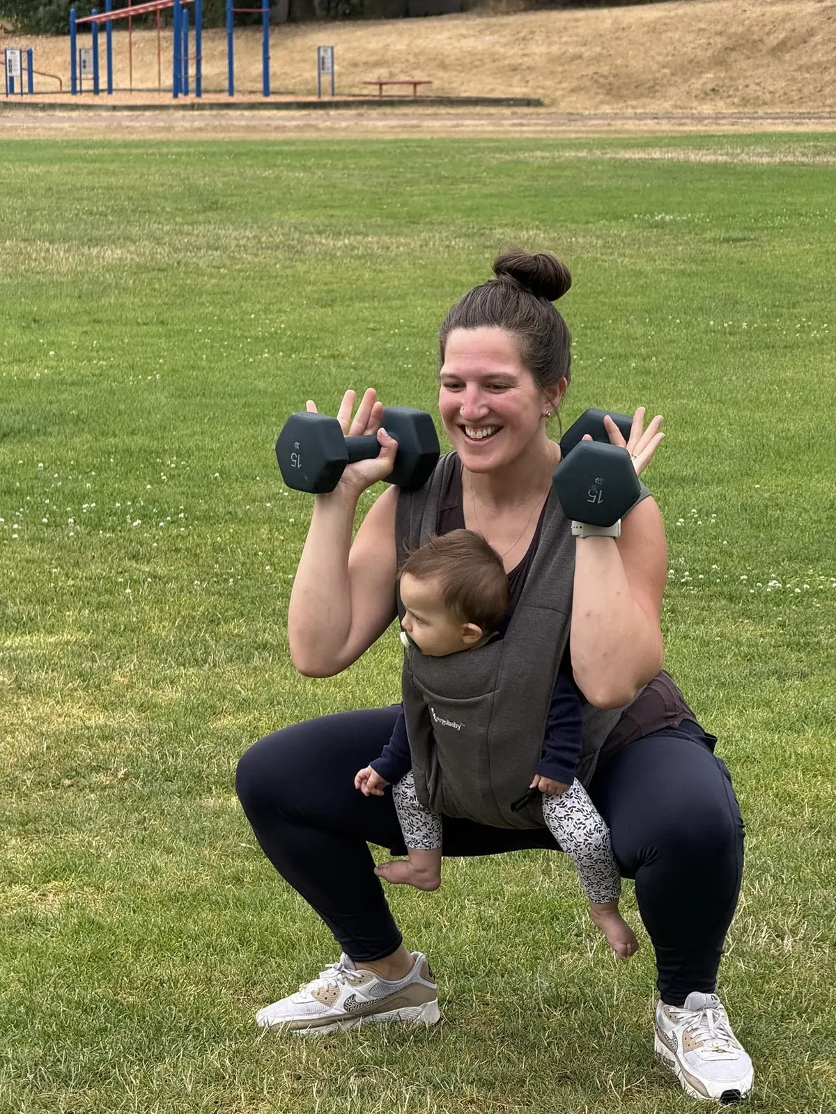 Mom squatting with dumbbells while baby rides in a front carrier during class